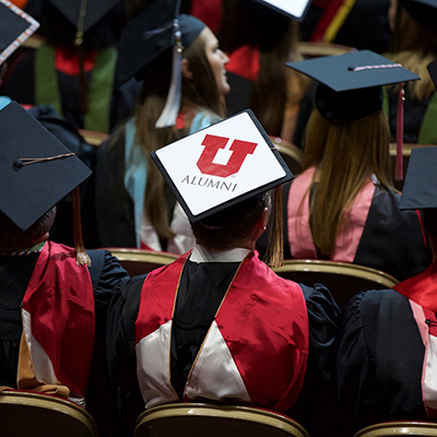 University of Utah graduates are photographed from above. The student in the center of the frame is wearing a cap adorned with a red block U above the word 